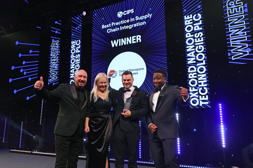 Image of two members of the Oxford Nanopore Technologies team on stage celebrating their award win holding the award. On either side are the host of the event (right) and a member of the CIPS team (left). The four people are standing on stage in front of blue graphics behind them on big screens saying the company's name, the award and 'Winner".